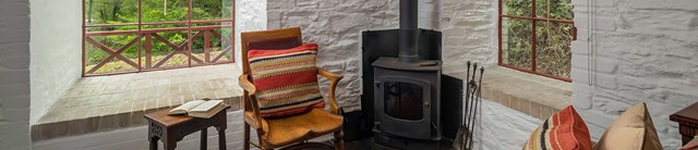 Danescombe Mine interior sitting room with wooden spoke-back chair, wood burning stove and views out of the pane-glass window to woodland